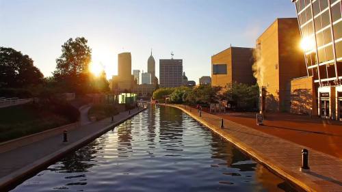 Sunset over a calm canal with city skyline reflections and buildings along the water's edge.