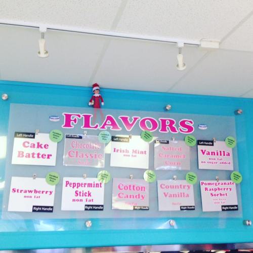 A colorful menu board displaying various ice cream flavors, with an elf figure perched on top.