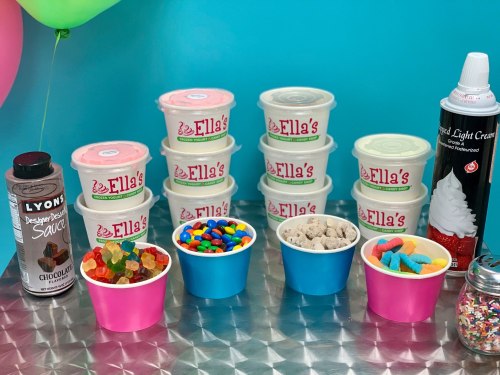 Colorful dessert setup featuring ice cream tubs, candy cups, and toppings on a shiny table with balloons in the background.