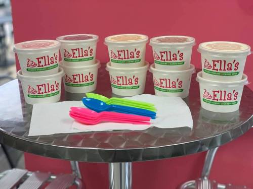 A display of Ella's frozen yogurt containers with colorful spoons on a table against a pink background.