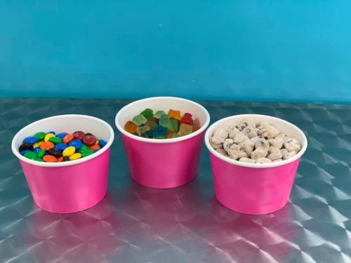 Three pink cups filled with colorful candies: one with chocolate candies, one with gummy candies, and one with cookie dough bites.