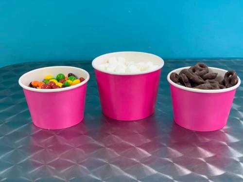 Three pink cups on a silver surface: one with colorful candy, one with marshmallows, and one with chocolate pretzels.
