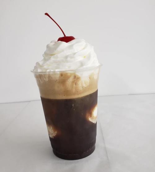 A clear cup filled with dark soda, topped with whipped cream and a cherry.