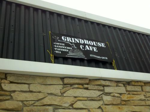 Sign for Grindhouse Cafe featuring coffee, sandwiches, pastries, and tea, mounted on a stone wall.