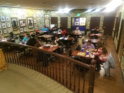 A busy room with people sitting at tables, engaged in activities, surrounded by framed photos on the walls.