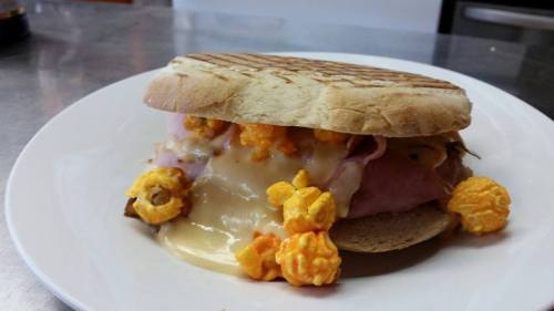 A sandwich with ham, melted cheese, and cheese popcorn, served on toasted bread on a white plate.