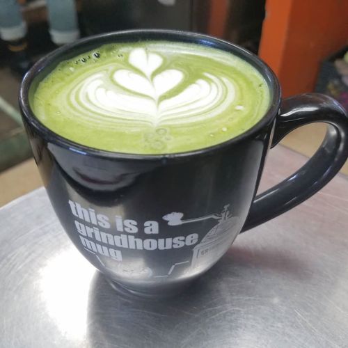A black mug with a heart-shaped latte art design, sitting on a metal surface. The mug reads "this is a grindhouse mug."