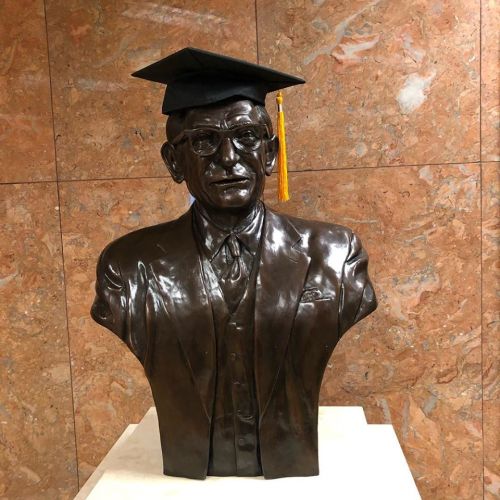 A bronze bust of a man wearing glasses and a graduation cap, set against a textured stone background.