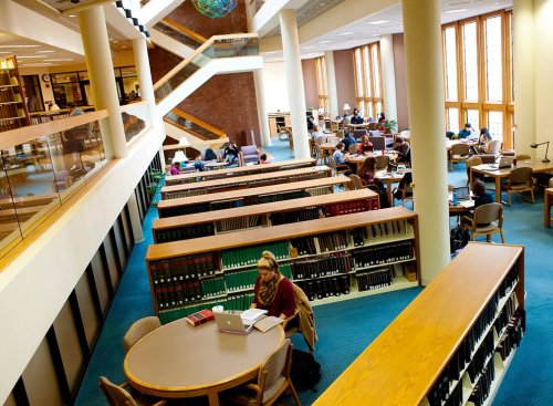 A spacious library with students studying at tables and shelves filled with books, featuring large windows and modern architecture.