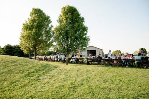 A long table set for a gathering outdoors, surrounded by trees and a grassy hill, with people enjoying a meal.