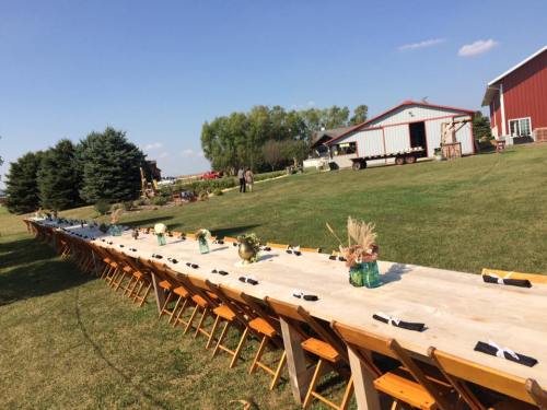 A long outdoor dining table set with black napkins, surrounded by greenery and rustic buildings under a clear blue sky.
