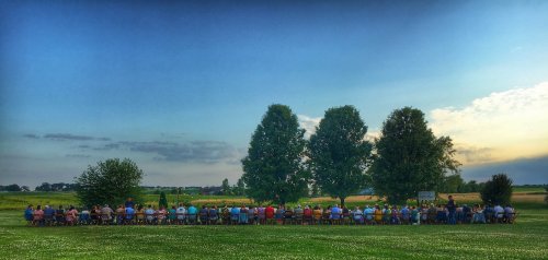 A large group of people seated at a long table outdoors, with trees and fields in the background under a blue sky.
