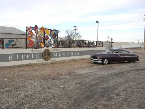 A vintage car parked near a colorful mural at the entrance of America's Hippie Memorial.