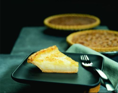 A slice of creamy pie on a black plate, with two whole pies in the background. A fork rests beside the plate.