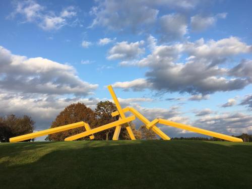 A large yellow sculpture made of angled beams on a grassy hill, with trees and a cloudy sky in the background.