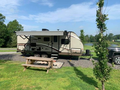 A camper trailer parked near a picnic table and a pond, surrounded by green grass and trees under a blue sky.