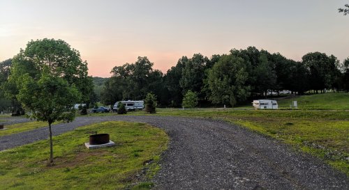 A gravel path winds through a campsite with trees, RVs, and a sunset sky in the background.