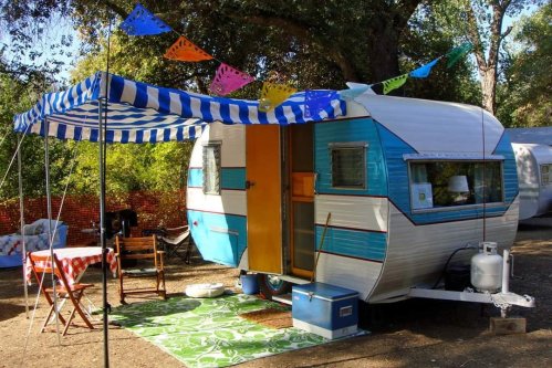 A vintage camper with a blue and white exterior, set up with a striped awning, colorful banners, and outdoor seating.