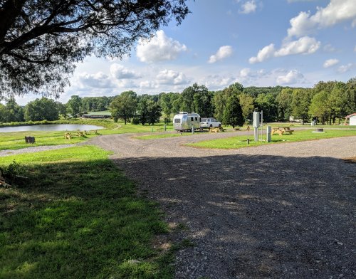 A gravel path leads to a campsite with an Airstream trailer, surrounded by green grass and trees near a pond.
