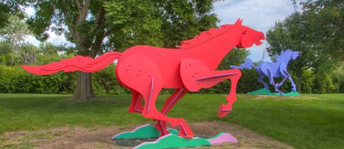Two colorful horse sculptures, one red and one blue, are displayed in a grassy area surrounded by trees.