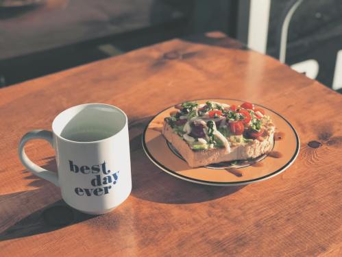 A mug that says "best day ever" next to a plate of colorful avocado toast on a wooden table.