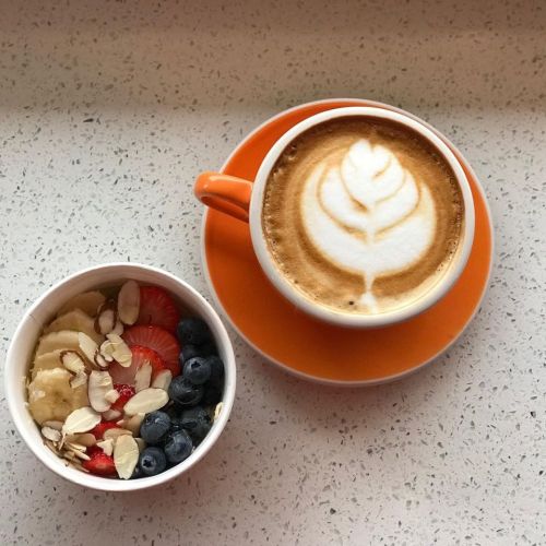 A cup of latte with a leaf design, next to a bowl of mixed berries, banana slices, and almonds on a speckled surface.