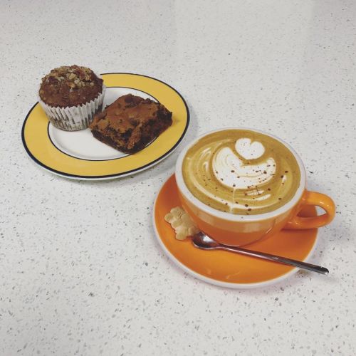 A vibrant orange cup of latte art sits beside a plate with a muffin and a brownie on a white countertop.