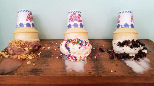 Three ice cream cones on a wooden surface, topped with chocolate, sprinkles, and crushed cookies.