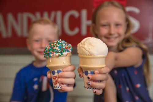 Two children hold up ice cream cones, one with colorful sprinkles and the other plain, smiling at the camera.