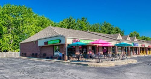 A brick storefront with colorful signs, outdoor seating, and umbrellas, surrounded by trees under a clear blue sky.