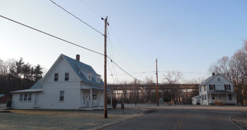 Two white houses at a road intersection, with power lines and a bridge in the background under a clear blue sky.