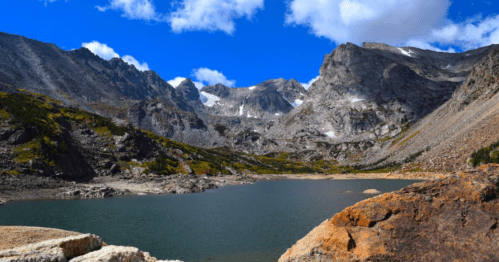 A serene mountain landscape featuring a clear lake surrounded by rugged peaks and a bright blue sky with clouds.