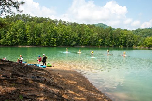 A serene lake scene with people paddleboarding and others on the shore, surrounded by lush green trees and mountains.