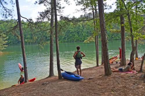 A serene lakeside scene with kayakers, trees, and people relaxing on the shore.