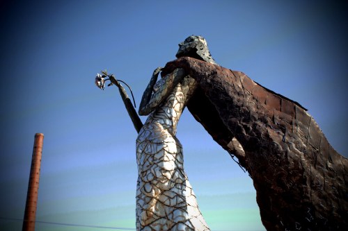 A large, abstract sculpture of a figure holding a flower, draped in a flowing cloak against a clear blue sky.