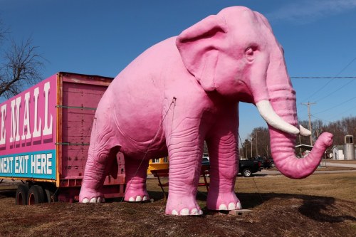 A large pink elephant statue stands beside a colorful trailer, set against a clear blue sky.