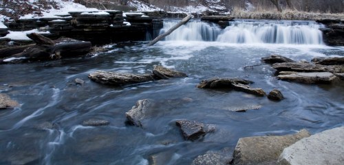 A serene river scene with flowing water over rocks, surrounded by snow and bare trees in a tranquil landscape.