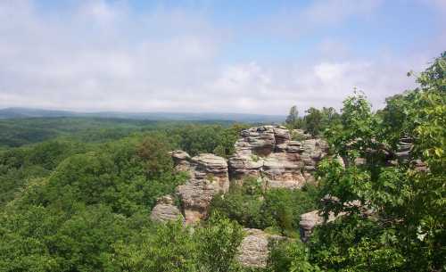 A scenic view of rocky formations surrounded by lush green trees and a cloudy sky in the background.