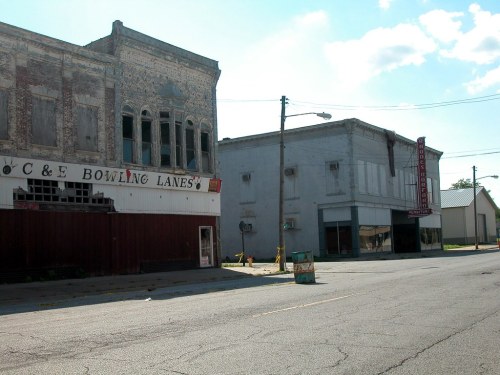 Abandoned buildings line a quiet street, featuring a vintage bowling alley and a closed storefront under a blue sky.