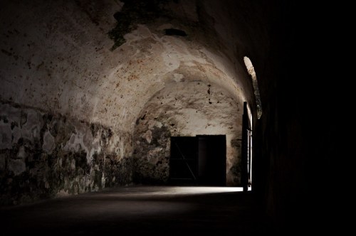 A dimly lit, empty stone corridor with arched ceilings and a dark doorway at the end.