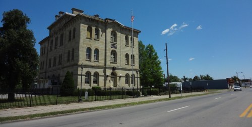 Historic stone building with a flag, surrounded by trees and a fence, alongside a road under a clear blue sky.