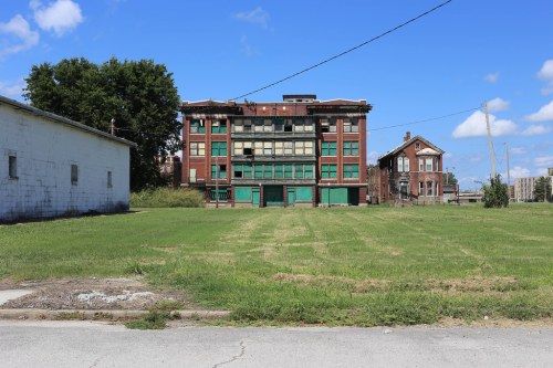 Abandoned brick building with broken windows, surrounded by overgrown grass and a clear blue sky.