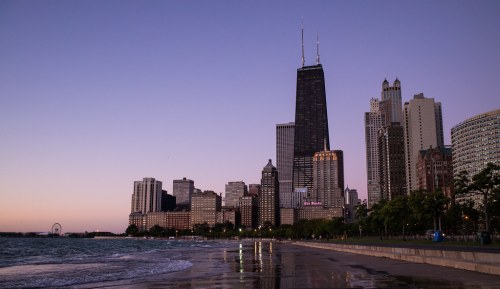 Chicago skyline at dusk, featuring the John Hancock Center, with a sandy beach and calm lake in the foreground.