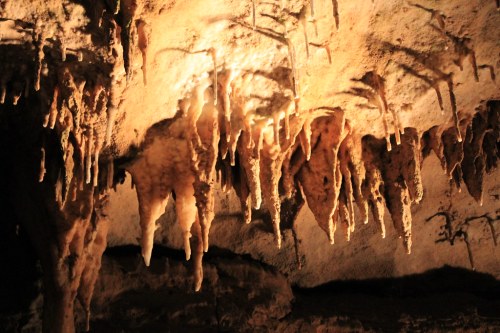 Stalactites hang from a cave ceiling, illuminated by warm light, creating a dramatic underground scene.