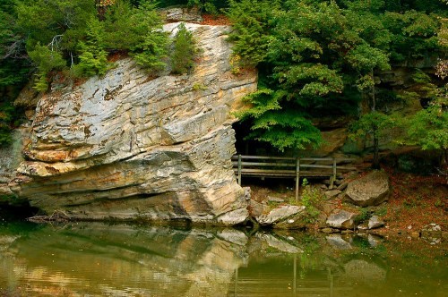 A rocky cliff beside a calm lake, with lush greenery and a wooden bridge leading to a small cave entrance.