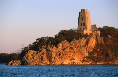 A stone tower sits atop a rocky cliff by a calm lake, illuminated by the warm glow of sunset.