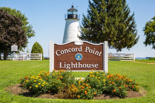 A sign for Concord Point Lighthouse in front of a white lighthouse, surrounded by green grass and colorful flowers.