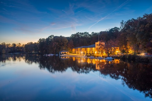 A serene lakeside view at dusk, featuring a warmly lit building reflecting on calm waters surrounded by trees.