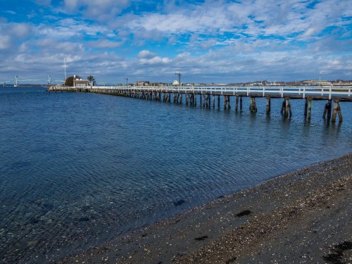 A wooden pier extends over calm water, with a sandy shore and cloudy blue sky in the background.