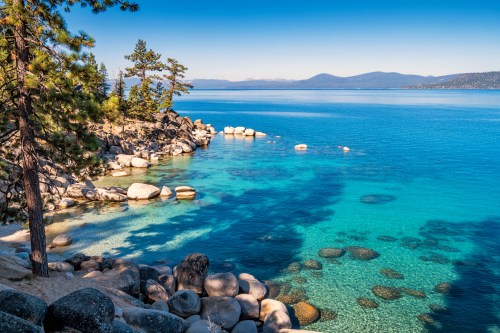 A serene lakeside view with clear blue water, rocky shores, and distant mountains under a bright blue sky.